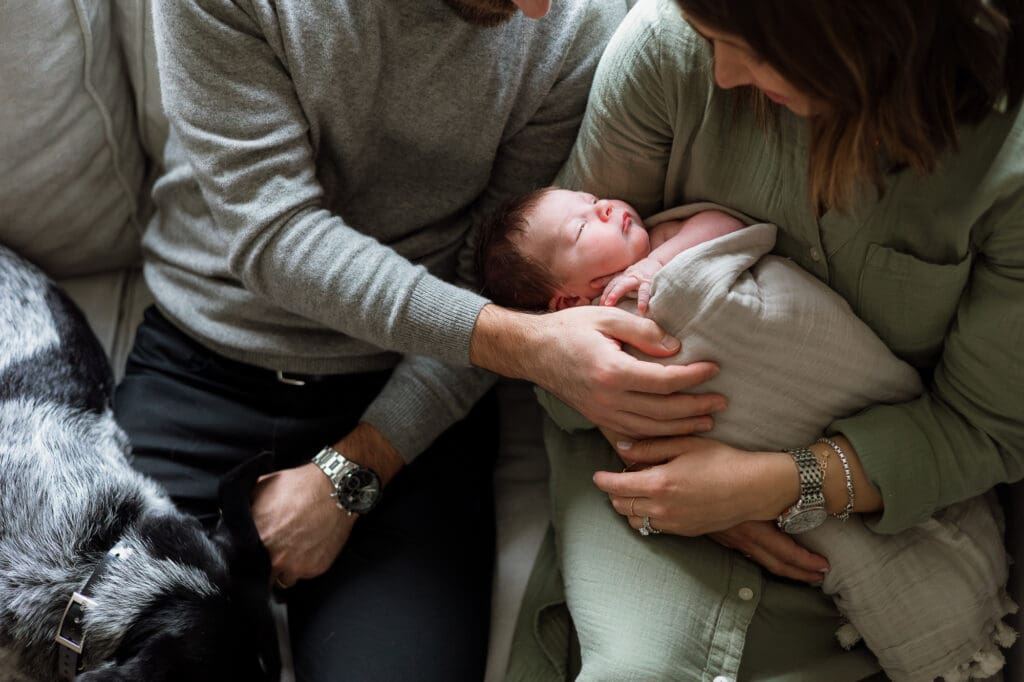 Mom and dad holding swaddled baby on couch with dog
