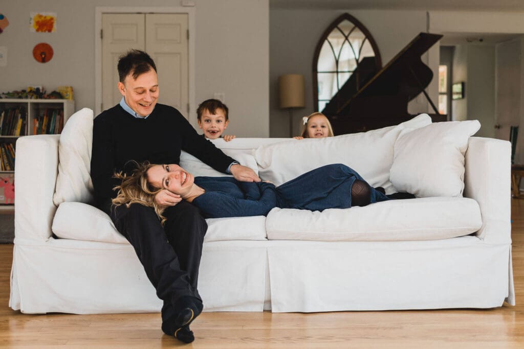 Mom and dad snuggling on couch in their home while kids peek over the back.