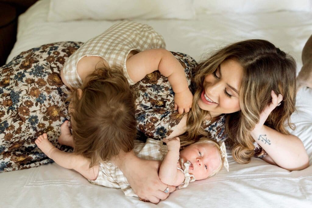A mother lounges with her newborn and toddler on the bed—capturing the chaos and connection of life during the newborn stage.
